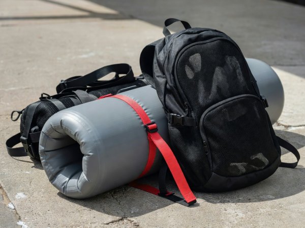Close-up photograph of a tightly rolled inflatable paddleboard secured with a crimson compression strap beside a ventilated black mesh stowage backpack